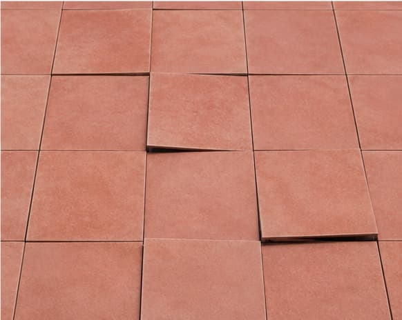 Terracotta floor tiles viewed from above, most flush and flat, some corners quietly lifted out of place.